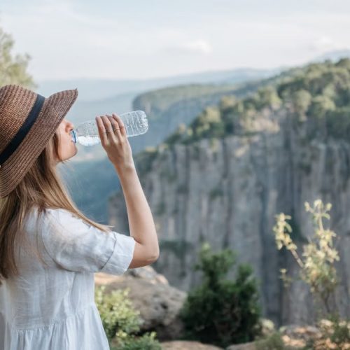 Woman wearing a sun hat drinks water while enjoying a scenic canyon view amidst nature.